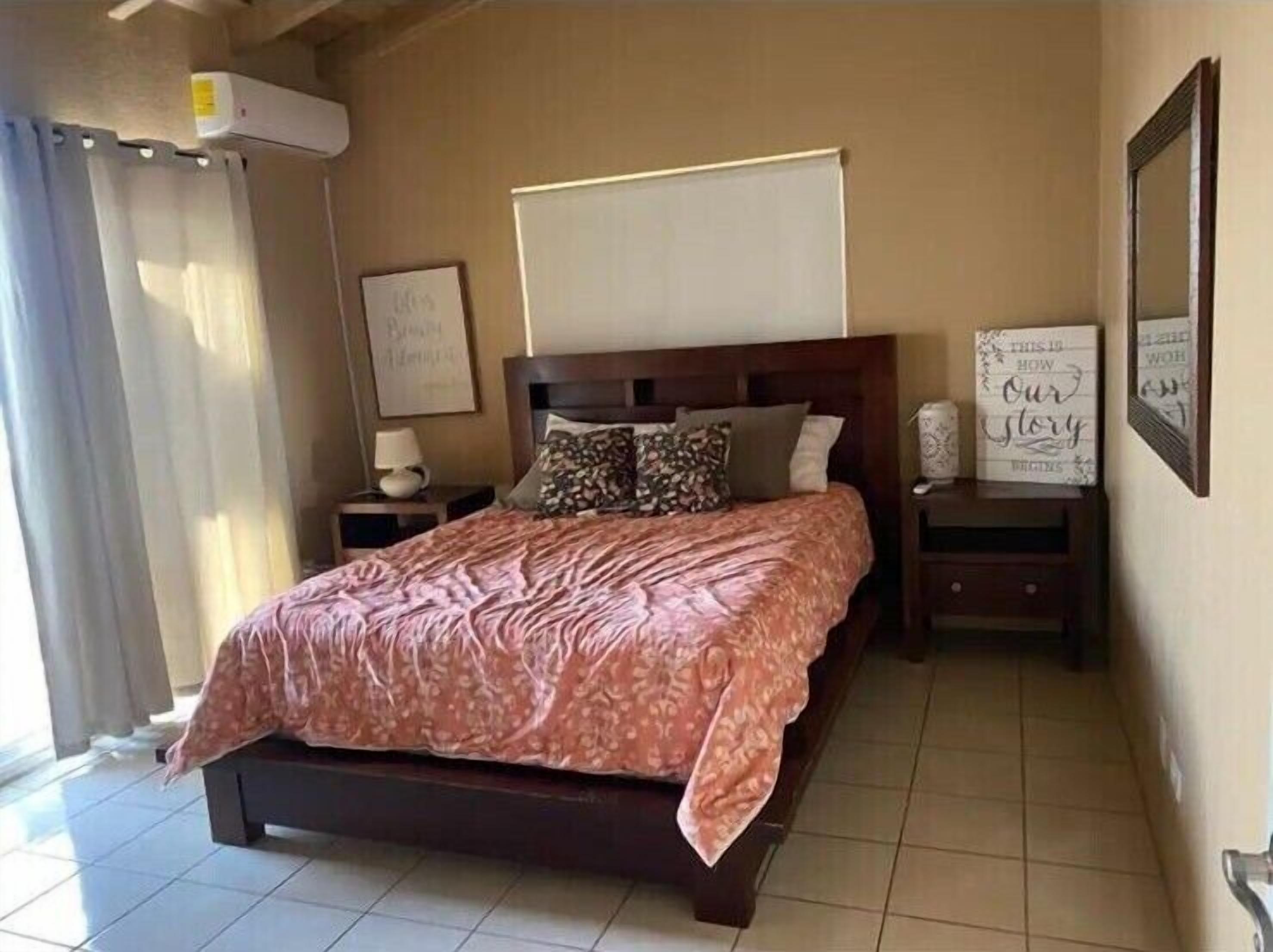 A bedroom with a large bed, a nightstand with a lamp, a wall-mounted air conditioner, and framed inspirational quotes.