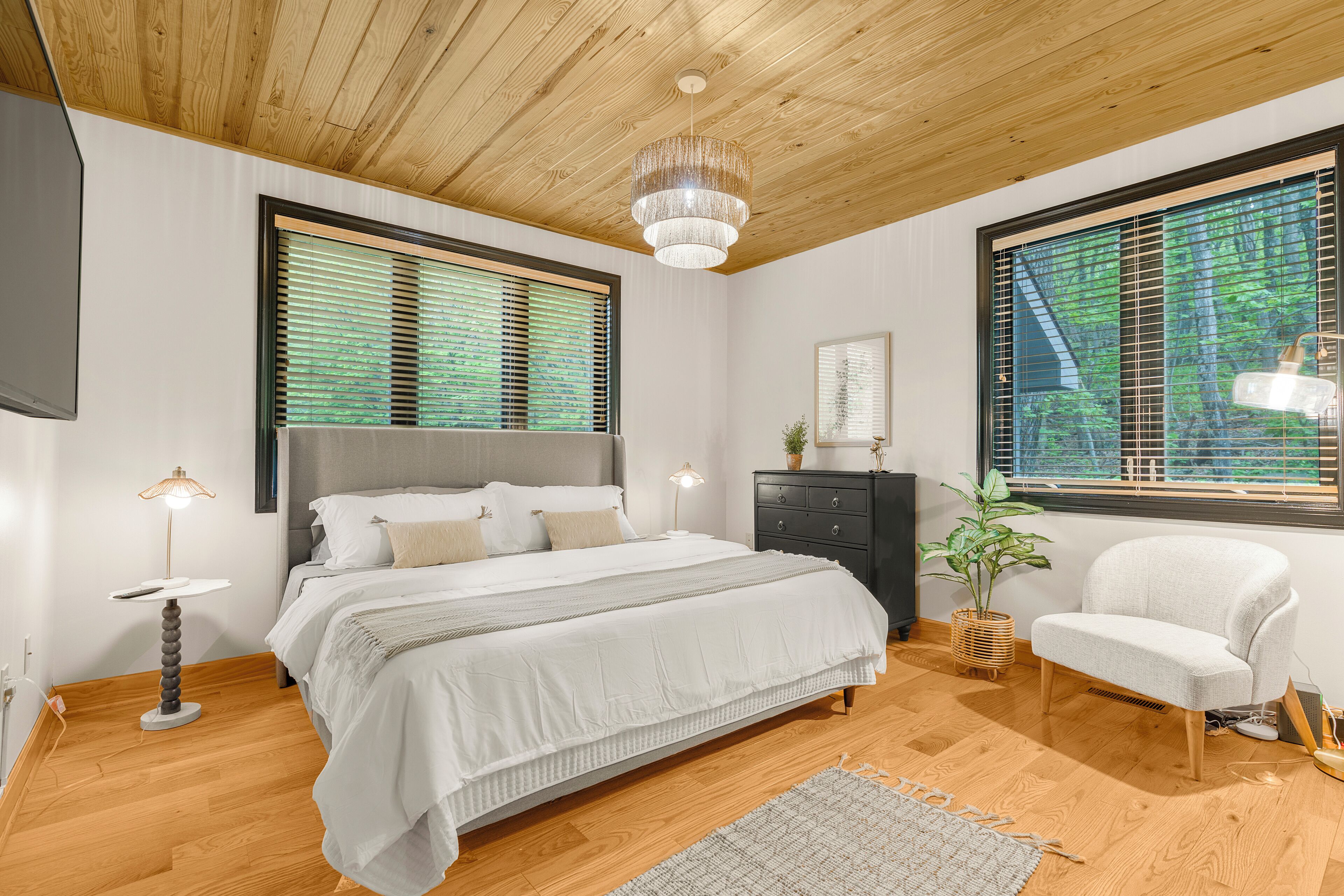 A bedroom with a wooden ceiling, a bed with white bedding, a bedside table with a lamp, a chair, and a window with blinds.