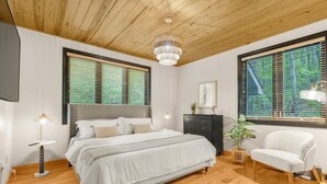 Une chambre avec un plafond en bois, un lit avec une literie blanche, une table de chevet avec une lampe, une chaise et une fenêtre avec des stores.