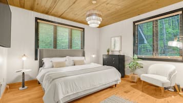 A bedroom with a wooden ceiling, a bed with white bedding, a bedside table with a lamp, a chair, and a window with blinds.