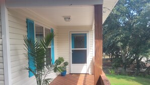 Un porche cubierto con una terraza de madera, una planta en maceta y una puerta con ventanal.