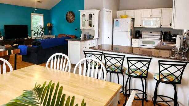 Una cocina moderna con armarios blancos, un comedor con mesa de madera y sillas blancas, y una sala de estar con una pared de acento azul y un ventilador de techo.