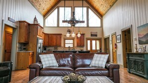 A spacious living room with a high ceiling, wooden ceiling beams, a brown leather sofa, a coffee table with decorative items, and a kitchen in the background. - Huge Decks, Hot Tub, Fire Pit & Dog Friendly. Close to Boone and Banner Elk. (Seven Devils)