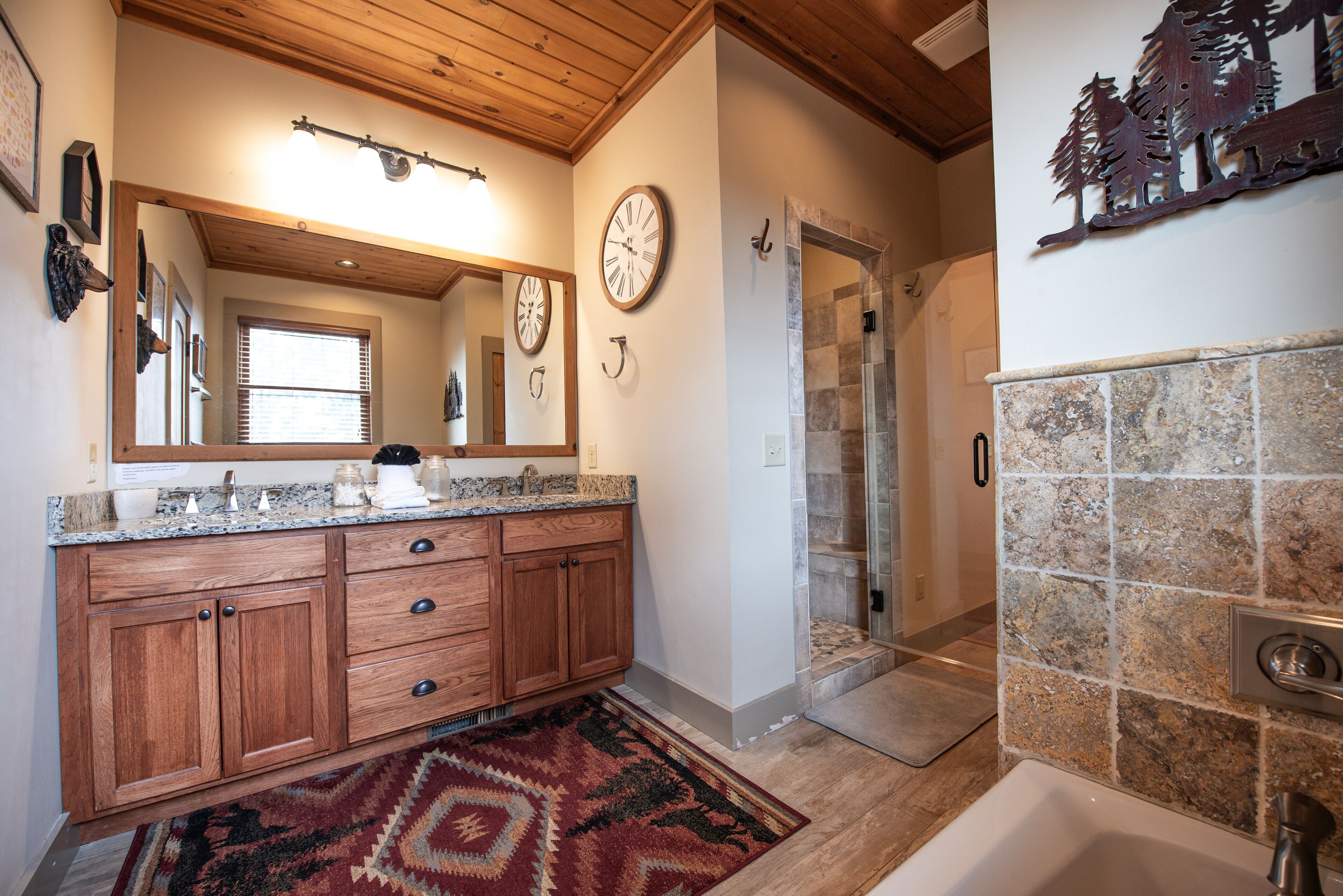 Une salle de bain avec des meubles en bois, un comptoir en granit, un grand miroir, une douche et un mur recouvert de carreaux de pierre.