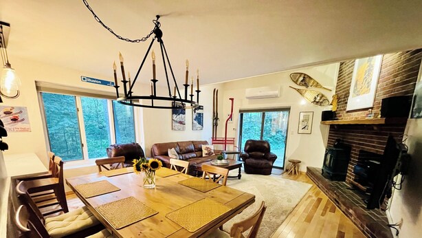 A spacious living room with a wooden dining table, comfortable seating, a fireplace, and large windows offering a view of the outdoors.
