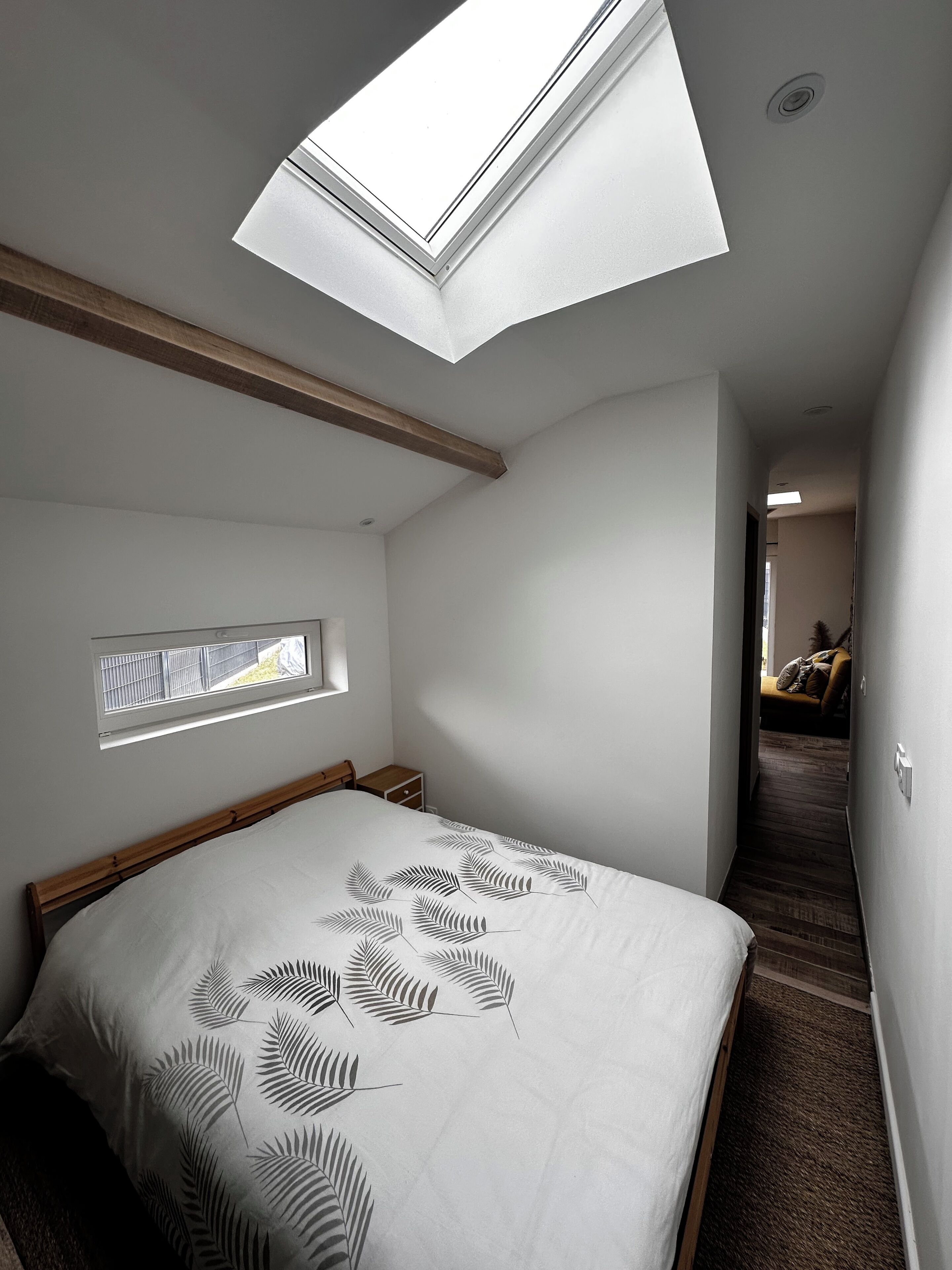 A bedroom with a skylight, a small window, a bed with a leaf-patterned bedspread, a wooden headboard, and a hallway leading to another room.