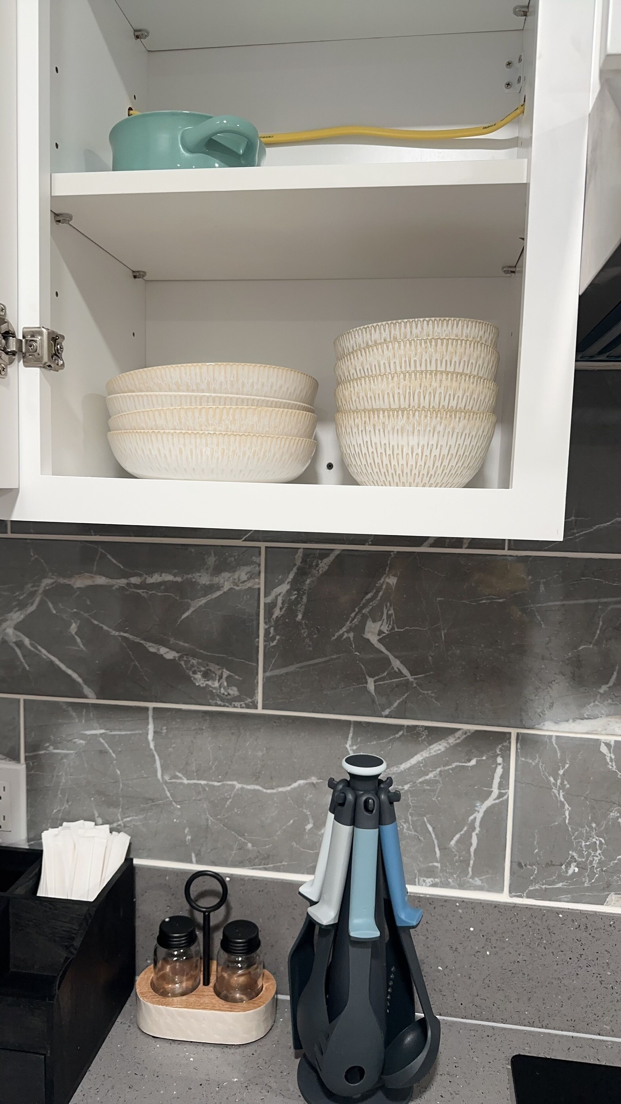 A kitchen with a marble backsplash, a white cabinet with two woven baskets, a yellow cable, and a kitchen utensil set on the counter.