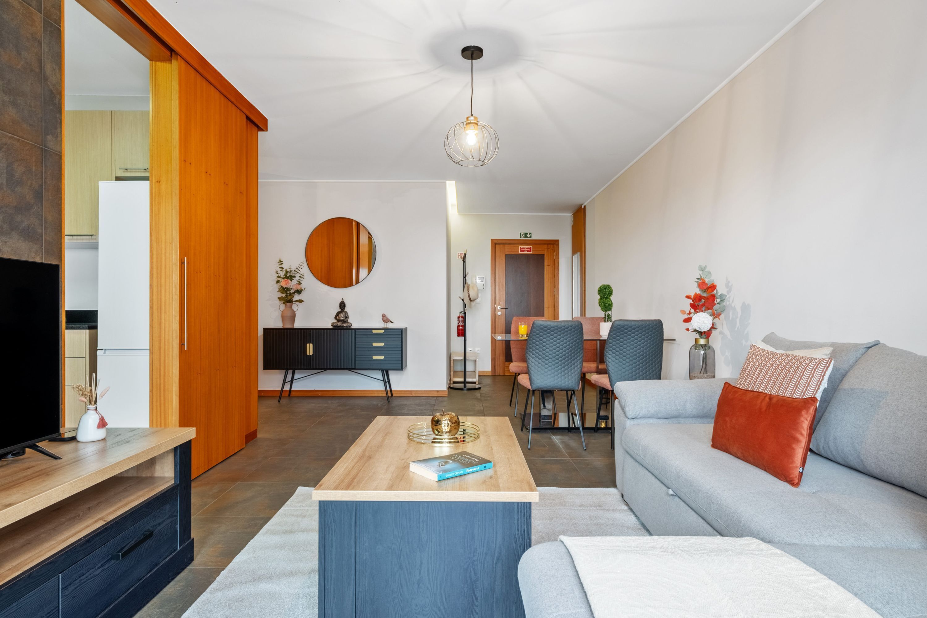 A modern living room with a sofa, coffee table, and TV stand. There is a dining area with chairs and a table, and a kitchen in the background.