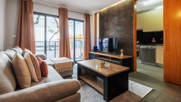 A modern living room with a sofa, coffee table, and TV. It features a large window with curtains, a kitchen area with cabinets and appliances, and a sliding door leading to an outdoor space. - Urban Stay 1A by Homie (Funchal)