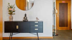 A modern room with a dark blue cabinet, a round mirror, a vase with flowers, a Buddha statue, and a bird figurine. There's a wooden door with a sign above it. - Urban Stay 1A by Homie (Funchal)