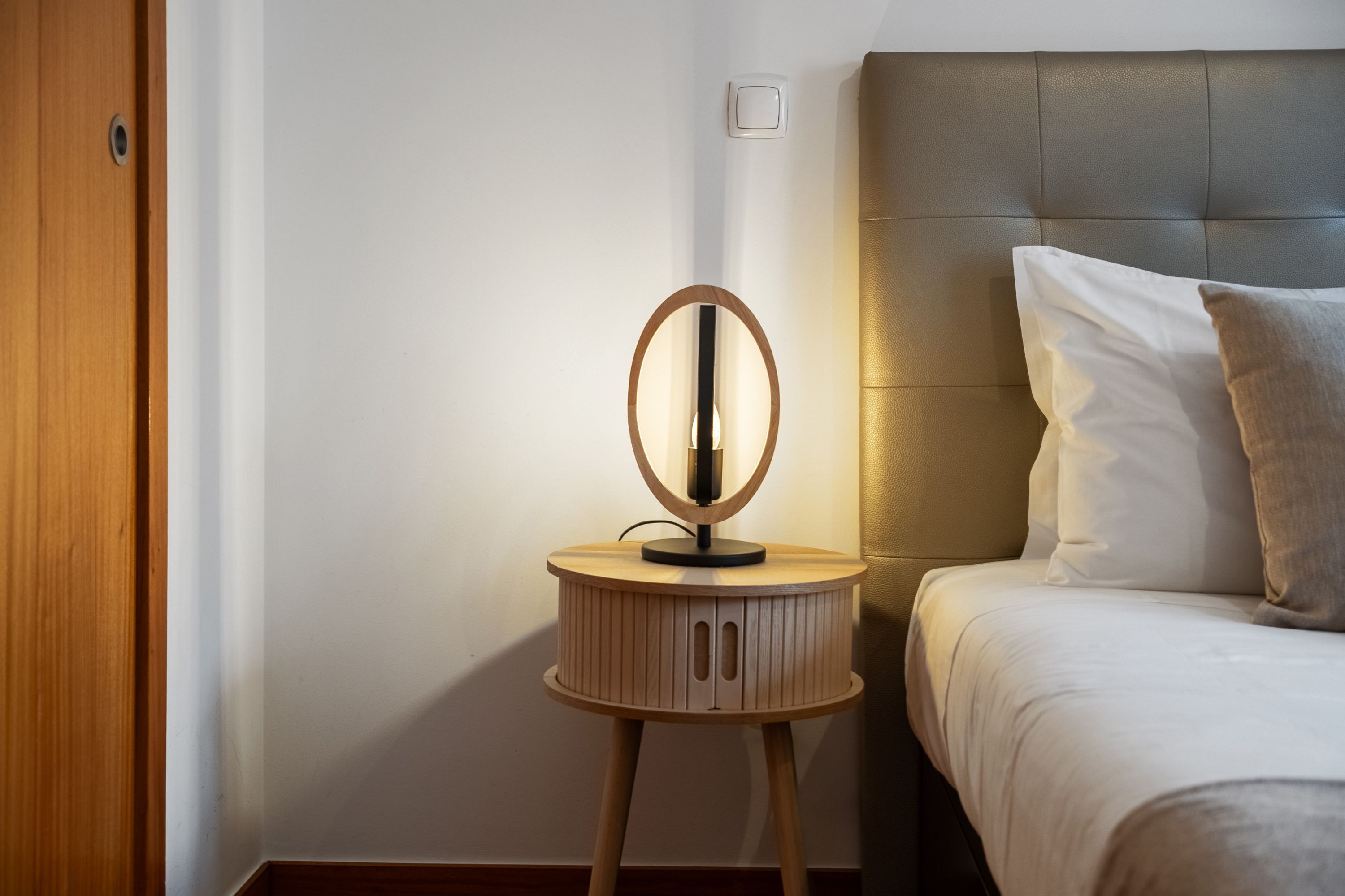 Un lit bien fait, avec une literie blanche et une tête de lit grise, une table de chevet en bois surmontée d’un miroir rond, et un interrupteur fixé au mur.