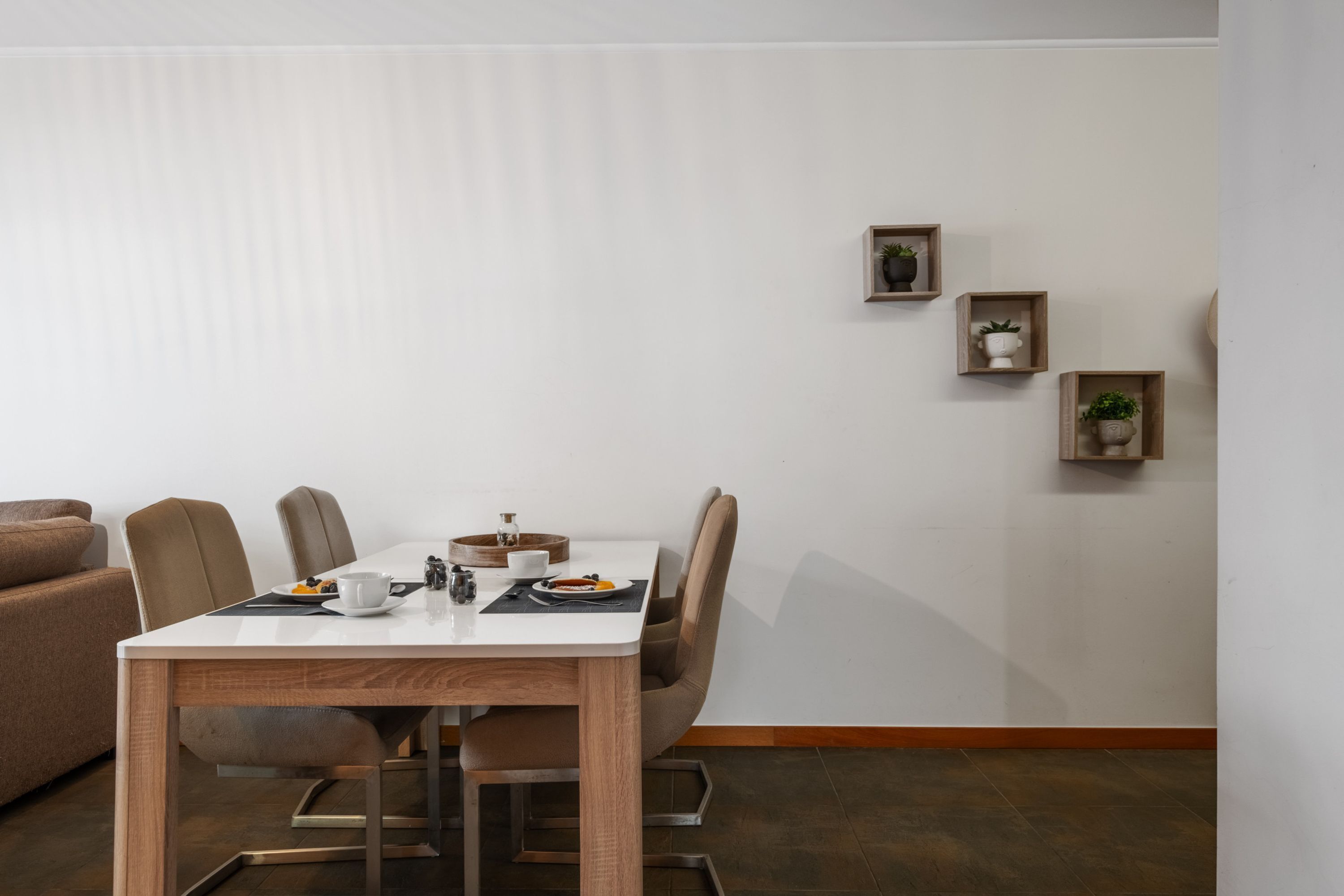 Een eetgedeelte met een witte tafel, bruine stoelen en een houten vloer. Er zijn drie kleine houten planken met planten aan de muur.