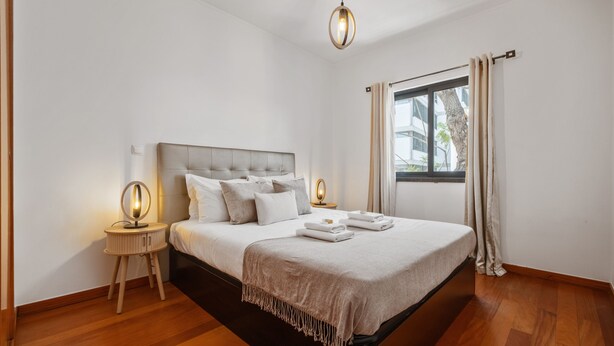 Un dormitorio moderno con una cama grande, lámparas de noche, una ventana con cortinas y vistas a edificios.