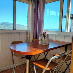 Un coin repas avec une table en bois et des chaises en métal, une fenêtre donnant sur des collines et un rideau.