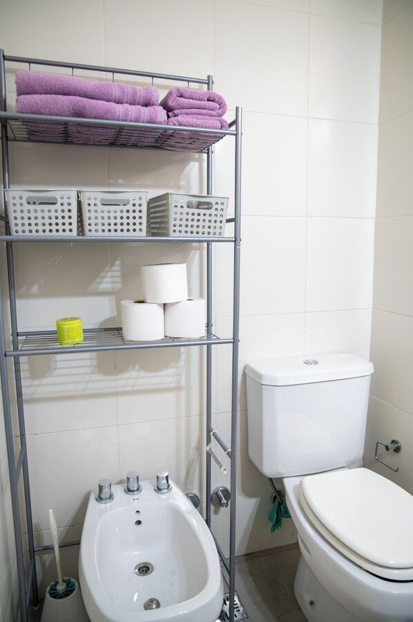 Un baño con inodoro blanco, un bidé, un estante con toallas, papel higiénico y una taza amarilla.