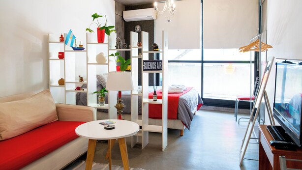 A modern room with a sofa, a small table, a TV, and a bed. There are shelves with various items and a window with a view. - Charming studio in beautiful Buenos Aires with AC, WiFi (Buenos Aires)