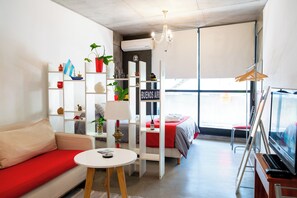 Una habitación moderna con sofá, una mesita, televisión y cama. Hay estantes con objetos diversos y una ventana con vistas.
