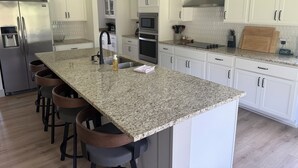 A modern kitchen with white cabinets, granite countertops, a stainless steel refrigerator, and a central island with bar stools. - Quiet 4BR 2.5BA Modern New Country Home in Weatherford (Weatherford)