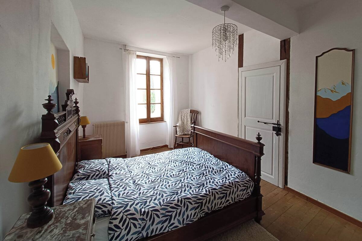 Um quarto com uma cama de madeira, um radiador, uma janela com cortinas, uma cadeira, uma porta e um quadro na parede.