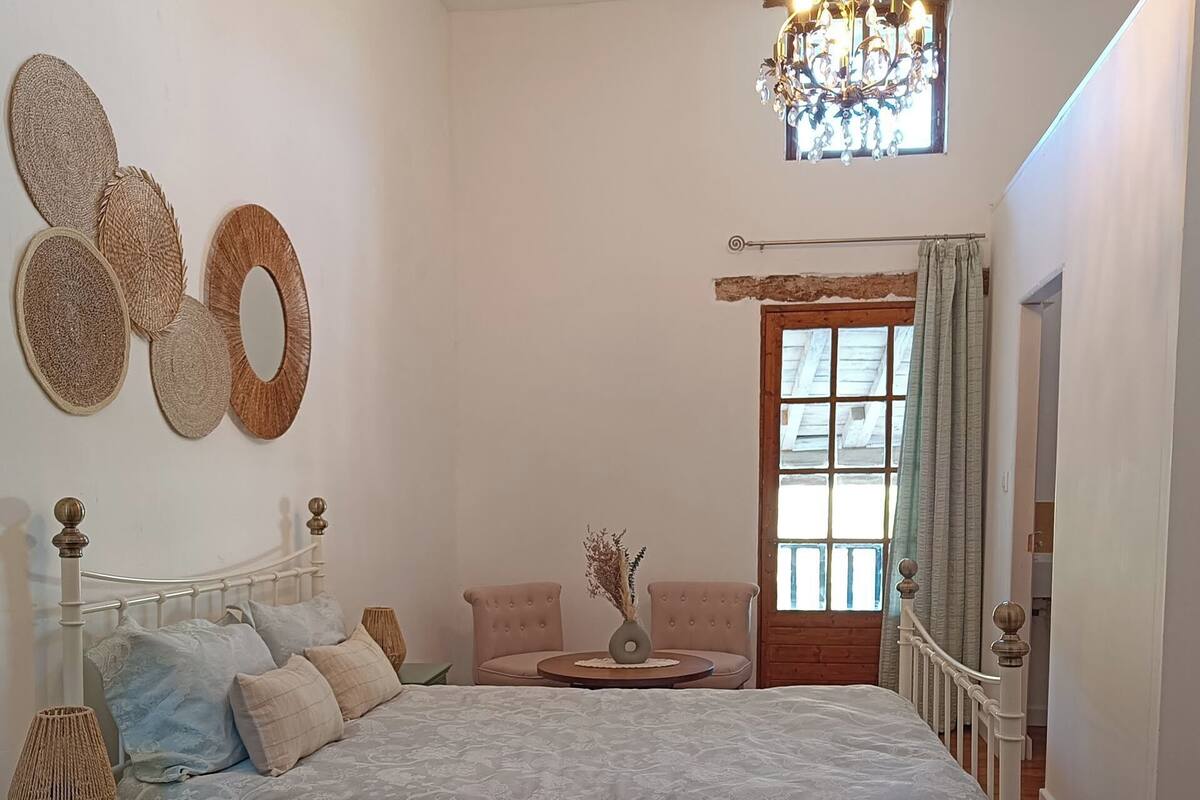 Quarto com cama, cadeira, mesinha, abajur e janela com cortinas.