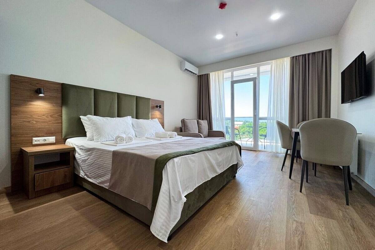 A modern hotel room with a large bed, a desk, a chair, and a balcony with a view.