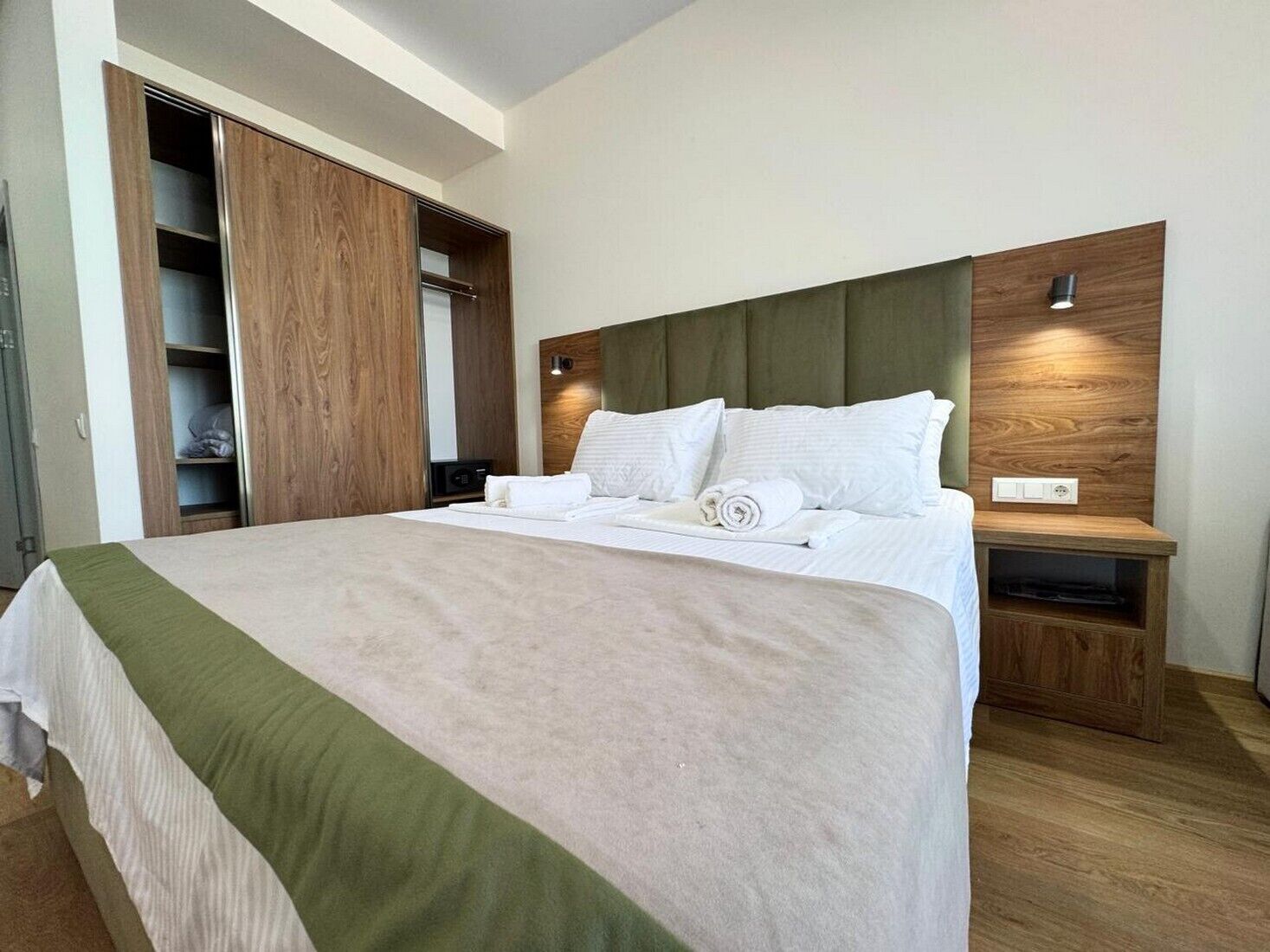 A modern hotel room with a large bed, wooden headboard, built-in wardrobe, bedside table, and a small TV.