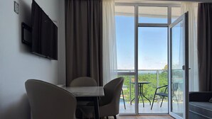 Een moderne kamer met een eettafel, twee stoelen, een bank, een aan de muur bevestigde televisie en een glazen schuifdeur die uitkomt op een balkon met uitzicht op groen.