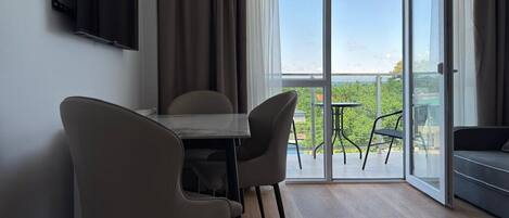 A modern room with a dining table, two chairs, a sofa, a TV mounted on the wall, and a sliding glass door leading to a balcony with a view of greenery.