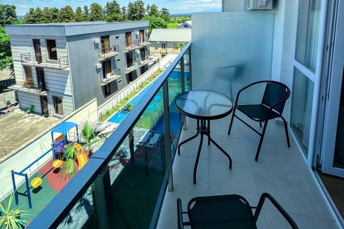 A balcony with two chairs, a glass balustrade, and a view of a swimming pool and buildings.