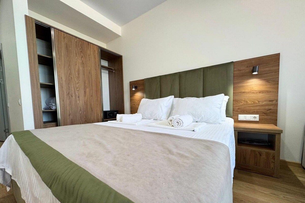 A modern hotel room with a large bed, wooden headboard, built-in wardrobe, bedside table, and a small TV.