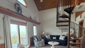 A spacious living area with a high ceiling, a ceiling fan, a staircase, and a view through large windows. - Charming Sound-front Captains Quarters in Frisco WiFi, streaming & perfect views (Frisco)