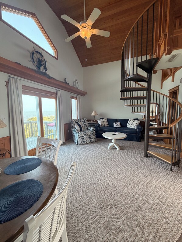 A spacious living area with a high ceiling, a ceiling fan, a staircase, and a view through large windows.