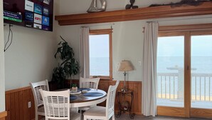 A dining area with a round table, white chairs, a potted plant, and a TV displaying various channels. - Charming Sound-front Captains Quarters in Frisco WiFi, streaming & perfect views (Frisco)