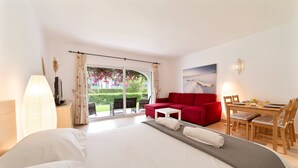 A hotel room with a large bed, a red sofa, a dining table with chairs, and a view of a garden through the sliding doors. - Apt. 77 - São Rafael by HelloVacations (Albufeira)