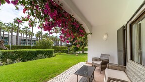A patio with wicker furniture, a tiled floor, and a view of a well-maintained lawn and palm trees. - Apt. 77 - São Rafael by HelloVacations (Albufeira)