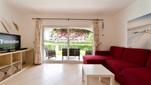 A living room with a red sofa, a white coffee table, a TV on a wooden stand, and a view of a garden through a glass door. - Apt. 77 - São Rafael by HelloVacations (Albufeira)