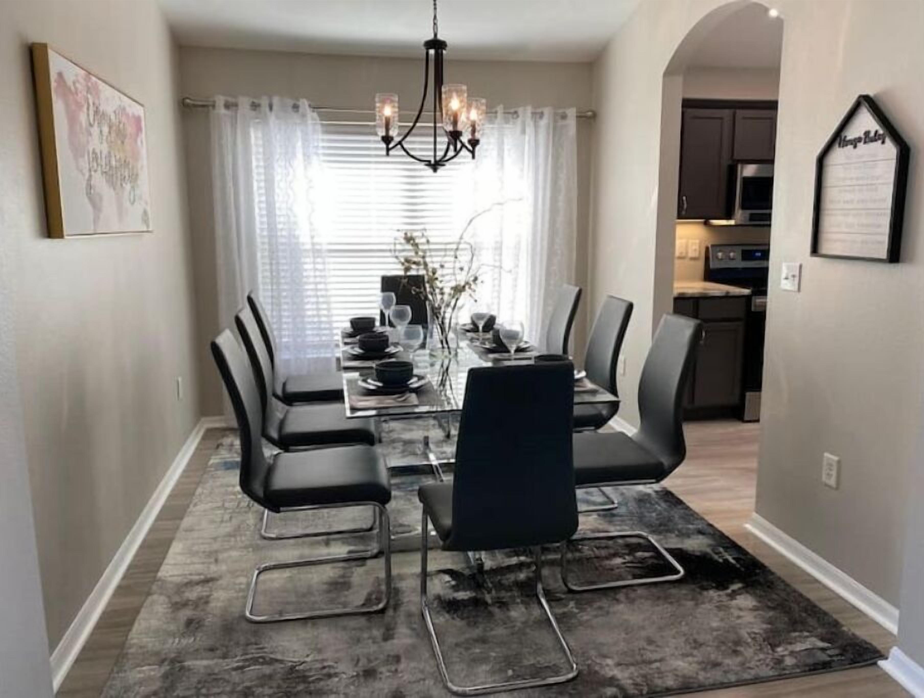A dining area with a glass table, black chairs, a chandelier, and a view of the kitchen.
