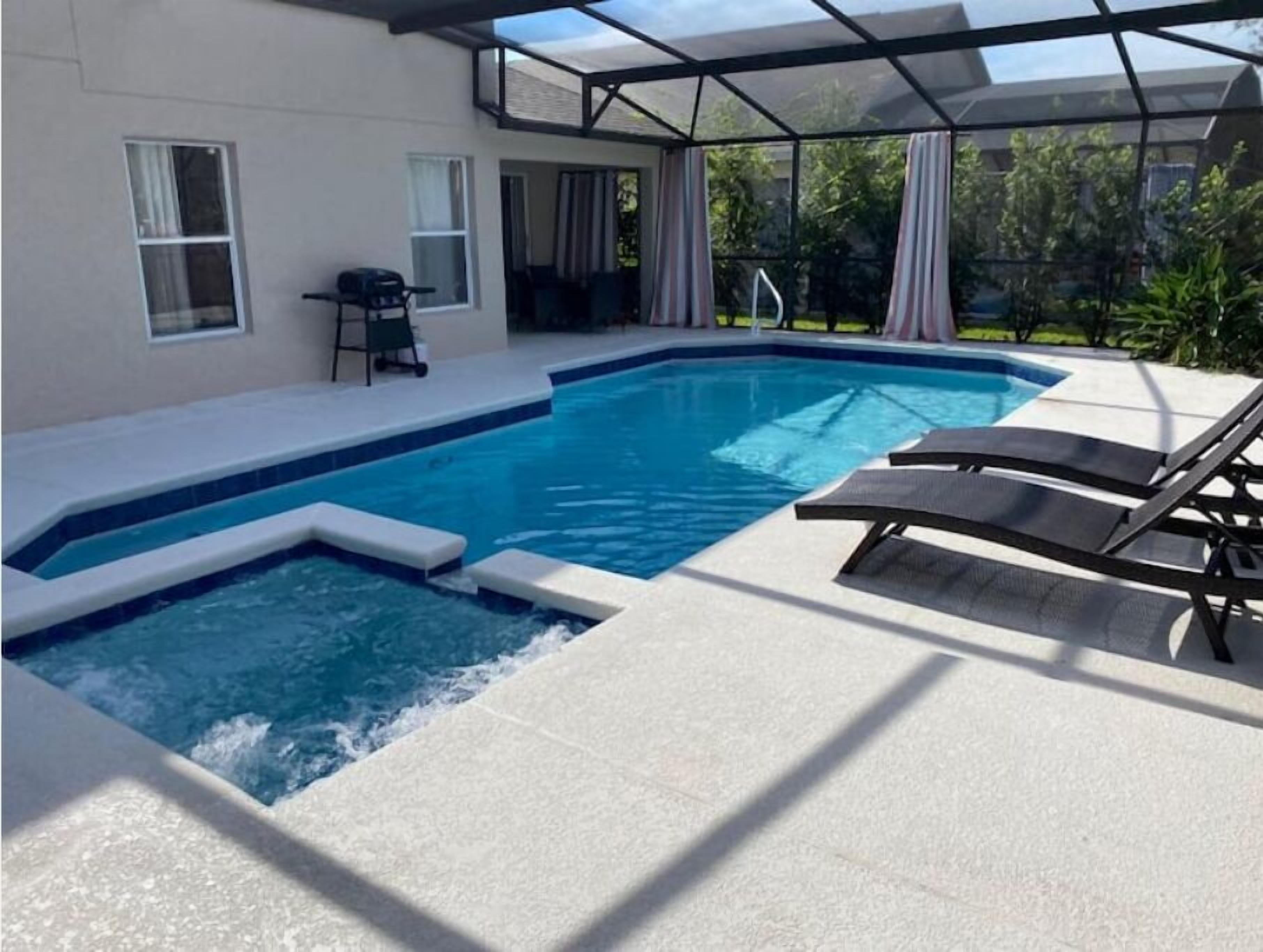 Une piscine avec un espace patio fermé par des parois de verre, une chaise longue et une table.
