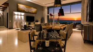 A modern living room with a large TV, a wooden dining table, and a view of the sunset through large windows. - Beachfront Apartment | 6 Bedrooms | Nuevo Vallarta Peninsula (Bucerías)