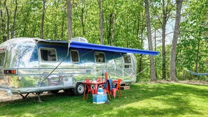 Un camping-car argenté est garé sur une pelouse, avec un auvent bleu, des chaises rouges et une glacière bleue.