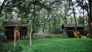 Kaksi puista mökkiä metsässä, niiden edessä on ruohikkoalue.