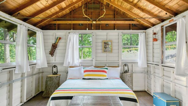 A cozy cabin bedroom with a wooden ceiling, a bed with striped bedding, and a small table with a lantern. - Sun View - Antique Scout Cabin Suite (Troy)