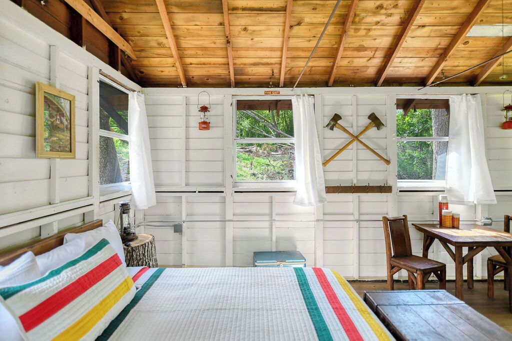 Una cabaña con techo de madera, paredes blancas, una cama con ropa de cama colorida, una mesa, sillas y dos ventanas con cortinas.