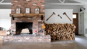 Une cheminée en briques avec une étagère, une pile de bois de chauffage et deux haches accrochées au mur.