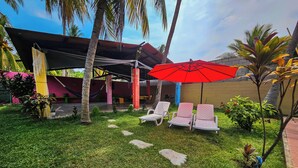 Un patio con tres sillones de descanso bajo un paraguas rojo, rodeado de palmeras y vegetación.