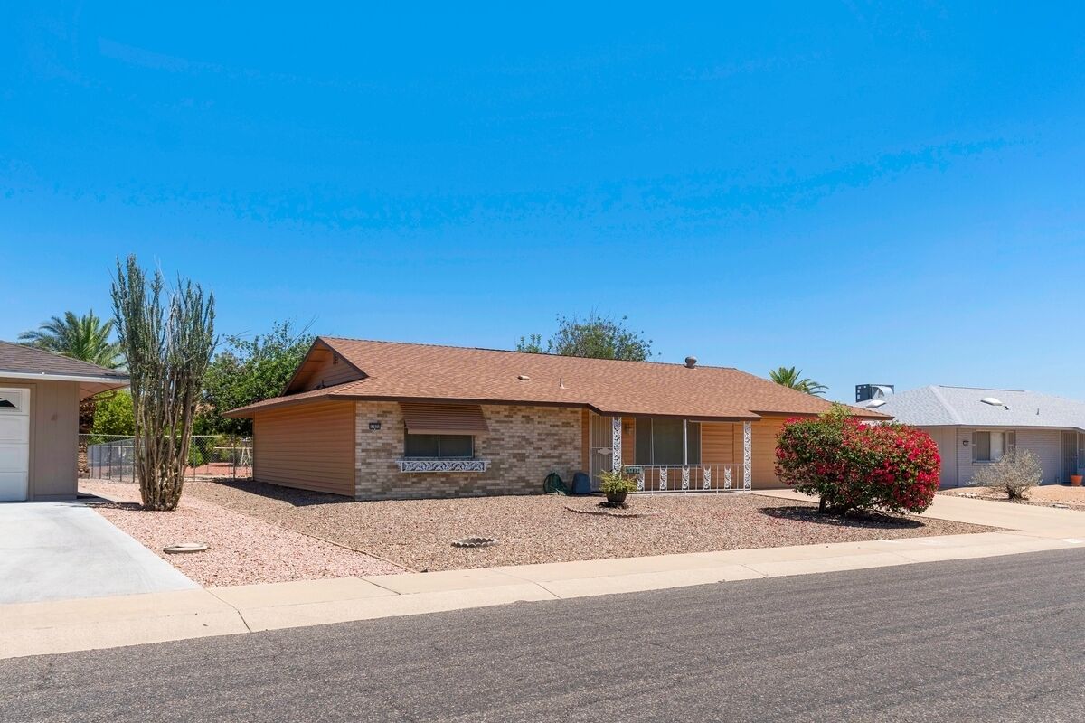 A single-story house with a stone facade, a brown roof, and a small front yard with a cactus and a flowering shrub.