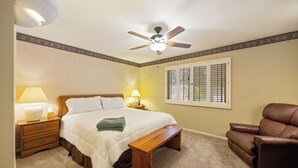 Ein Schlafzimmer mit einem Bett, Nachttischlampen, einem Deckenventilator, einem Fenster mit Jalousien, einem braunen Ledersessel und einer Holzbank.