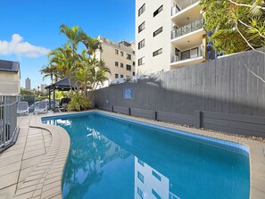 A swimming pool with a wooden fence, lounge chairs, and a view of a multi-story building.