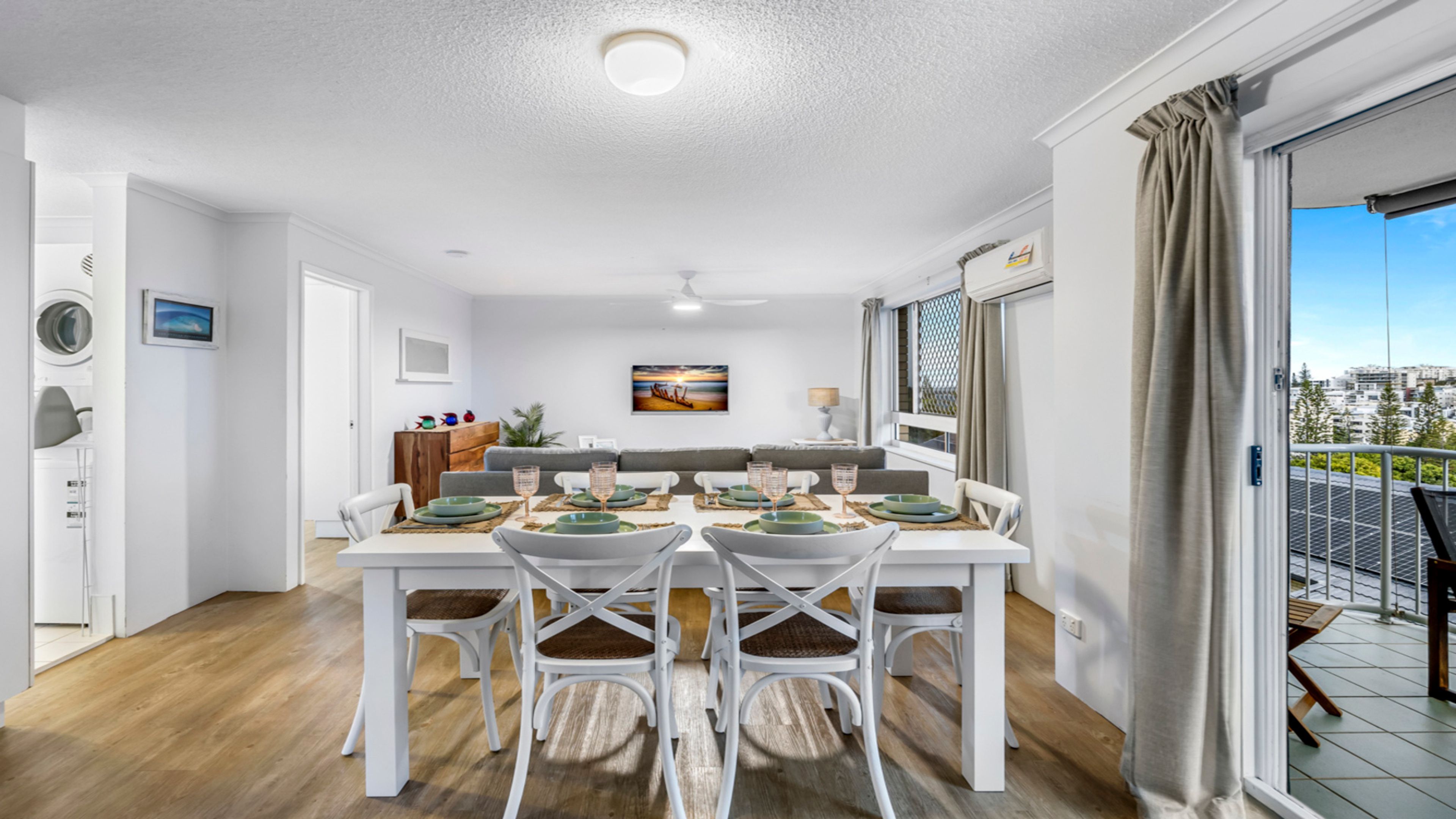 Zona de menjador amb una taula preparada per a quatre persones, envoltada d'unes cadires blanques, a un apartament modern amb vista a la ciutat.