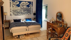 Ein Schlafzimmer mit zwei Betten, einem Holztisch, einem Stuhl, einem großen Wandteppich und einem Fenster mit Vorhängen.